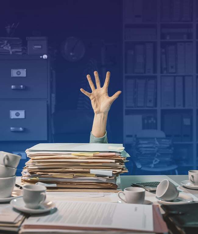 A hand reaching out from behind a tall stack of messy papers on a desk, symbolizing being overwhelmed by manual administrative tasks and paperwork.