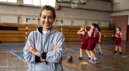 A female athlete smiling in a gymnasium with basketball players in the background, symbolizing efficiency in school sports.