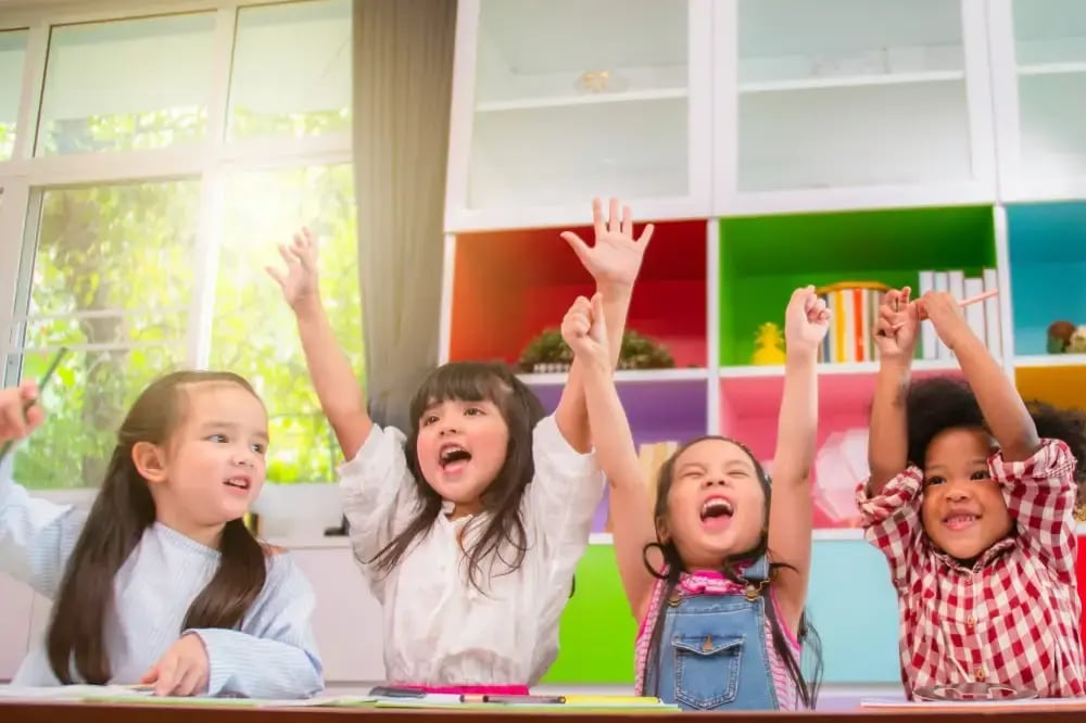 Four young children, smiling and raising their hands in a classroom.